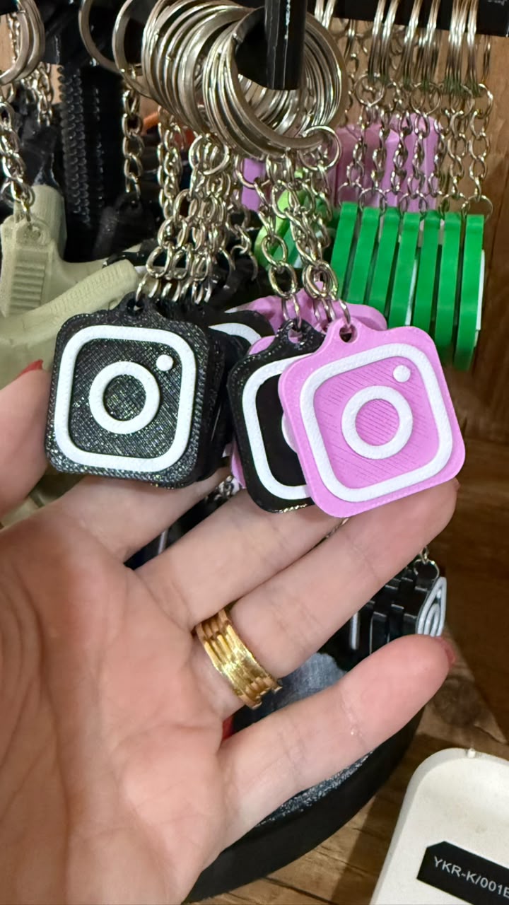 Mão segurando chaveiros com NFC em formato do ícone do Instagram, nas cores preta, branca e rosa, pendurados em um expositor.